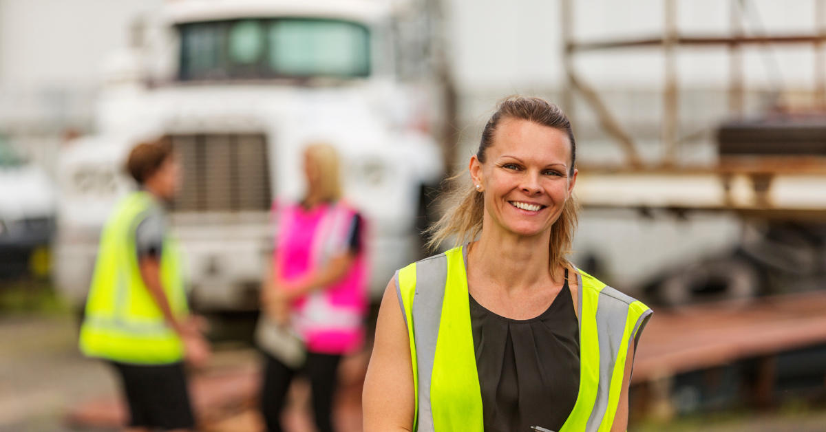 Woman in high vis clothing with a pen and notepad in front of two other workers in high vid clothing in front of a truck.