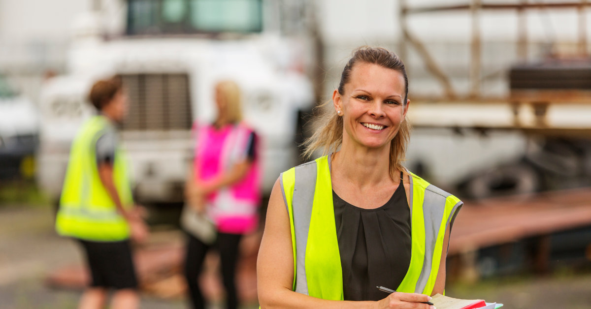 Woman in high vis clothing with a pen and notepad in front of two other workers in high vid clothing in front of a truck.